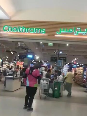 The video clip shows the entrance and interior of a Choithrams supermarket, located in Dubai, United Arab Emirates. The store's name is clearly visible above the entrance in illuminated green English letters, "Choithrams," alongside its Arabic script counterpart.

The scene depicts a busy retail environment with several people present. In the foreground, a person wearing a distinctive pink and black uniform, possibly a delivery driver, is seen from behind, pushing a shopping trolley into the store. Inside, shoppers are visible at various checkout counters and browsing the aisles. One man in a yellow shirt and grey pants walks past a checkout.

The store's interior is well-lit with numerous overhead lights. Checkout counters are visible, some with "CHECK-OUT" signs, and there are self-checkout kiosks on the right side of the frame. Shopping carts and green plastic baskets are scattered throughout the checkout area. The aisles appear fully stocked with various products, extending into the background. The overall impression is that of a modern, active grocery store during business hours.