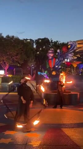 The video shows a fire performance taking place outdoors at dusk. Two individuals are performing, one in the foreground to the left, and another further back to the right. Both performers are dressed in dark clothing. The performer on the left is holding what appears to be a fiery staff or hoop, manipulating it in front of them. The performer on the right is near a large, fiery structure, possibly a pyre, that is ablaze. Sparks and flames are visible, creating a dynamic and visually striking scene.

In the background, there are large, colorful, whimsical structures that resemble fantastical sea creatures or characters. One appears to be a purple and red ice cream cone-like object, and another is a blue structure with an octopus on top. Trees and buildings are visible behind these decorative elements, suggesting an urban or park-like setting. The sky is a twilight blue, indicating that the sun has set or is setting. Lights from the surrounding area, including some stage lighting on the left, contribute to the illuminated atmosphere. The overall mood seems energetic and theatrical, with the bright flames contrasting against the darkening sky. The ground appears to be paved, with some patterned markings in the foreground. The location context of Taihe District, China, implies this is likely a public performance or festival. No visible text is present in the video.