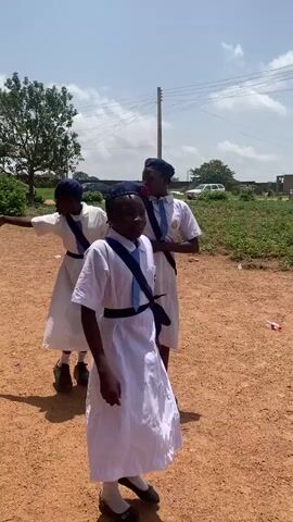 The video shows a group of young girls in white dresses and matching hats. Some have blue neckties and belts. They appear to be in a park or open field. One girl points in a direction while walking arm-in-arm with another. Another girl stands with her hands on her hips. A person in similar attire walks in the background. The ground is red dirt. In the distance, there are trees, a car, and power lines. The sky is blue with some clouds.