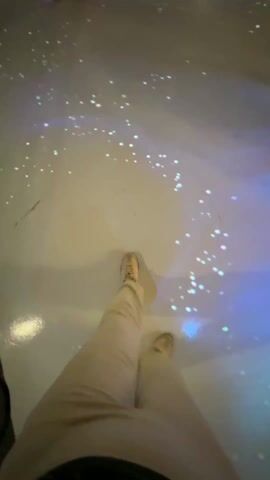 Here's a description of the video:

The video shows a first-person perspective of someone walking on a light-up floor with sparkling light patterns. Other people, including a child in pink pants and another wearing a dress, are walking around in the same area. The floor is smooth and light-colored, and the sparkling lights appear to be projected onto it. The person filming is wearing light-colored pants and shoes. The video focuses primarily on the floor and the movement of feet across it. There is some conversation heard in the background.