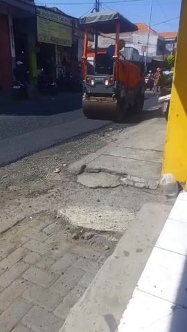 Here is a description of the video:

The video shows a road roller moving along a paved street. The road roller is orange and grey and has a protective frame around the operator's seat. It is labeled "HAMM" on the front. The road roller appears to be compacting freshly laid asphalt, visible as a dark strip in front of the roller. 

To the right of the road roller is a cracked and uneven sidewalk, composed of grey paving stones. In the background, various buildings are visible, including a shop with a sign reading "WARKOPNAS CAK MAN". People on motorcycles are seen driving on the street, and pedestrians are visible as well. The sky is clear and bright, suggesting a sunny day.