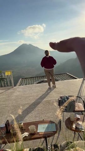 Here is a description of the video:

The video shows a person standing on a terrace overlooking a mountain range. The person is wearing a red jacket and tan pants, and they are facing away from the camera, looking at the mountain. The person holds up their phone and takes a photo of the mountain. In the foreground, there are two tables with coffee cups and a menu on them. There is also a backpack and a blanket on one of the chairs.  A hand is visible in the foreground, partially obscuring the view. The sky is blue, and there are a few clouds in the distance.