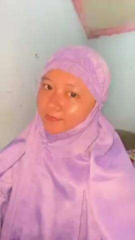 A woman wearing a light purple hijab is applying lip gloss. She holds a red-handled applicator to her lips and looks at the camera, turning her head slightly to the left. The background is a blue-green wall with a section of wood trim at the top.