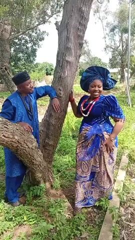 Two people dressed in traditional blue attire stand beside a tree. The man on the left leans against the tree and gestures with his hands. The woman on the right stands with her hand on the tree and smiles. The ground is covered with green grass and leaves. The background shows more trees and a light-colored building.