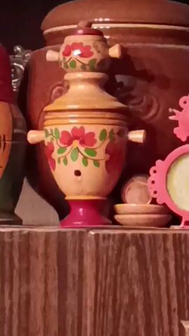 The video shows a shelf in a wooden cabinet. On the shelf, there are several decorative items. In the center, there is a wooden samovar-shaped toy, painted with red flowers and green leaves. To its left is a Russian nesting doll, painted in red and green. To the right of the samovar is a small pink clock with a round face. Further to the right, there is a small wooden house with a pitched roof. Behind the samovar is a large brown teapot with a lid. The shelf itself is made of dark wood and has a light-colored border. Below the shelf, the cabinet has two glass doors, revealing a collection of glassware, including glasses, cups, and saucers, as well as some decorative bowls. The background is dark.