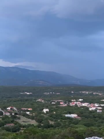 This is a landscape video of a rural village nestled in a valley, with mountains in the background. The scene is viewed from a high vantage point, offering a panoramic perspective. The village consists of numerous houses with red and white roofs scattered among lush green trees. The surrounding terrain is heavily forested, with a winding road visible in the distance.

The sky is overcast with dark, dramatic clouds, suggesting impending rain or a recent storm. The light is dim, indicating it is either early morning or late evening, likely dusk given the intensity of the clouds. The overall mood is serene and somewhat melancholic due to the weather.

There are no people or specific objects of focus, the emphasis is on the natural landscape and the settlement within it. The location is identified as Višnjevo, Montenegro. The video is approximately 3 seconds long and contains no audible sound or visible text.