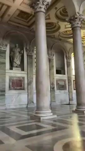 The video shows the interior of a large, ornate building, likely a church or historical site, with high ceilings adorned with decorative panels. There are large pillars throughout the space, and marble accents on the walls and floors. Windows with yellow stained glass are also visible. Several statues and framed artwork are displayed. Two people, an older man in a blue jacket and a woman, are seen walking through the area. The camera moves slightly, providing different views of the interior.