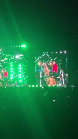The video shows two performers on a stage during a concert, viewed from the audience. The stage has a large screen with the performers' images, and lighting is used to enhance the performance. One performer is wearing a red jacket while the other is in a jacket with tan stripes. The audience is visible in the foreground, with many holding up their phones. Green lights illuminate the stage. The overall atmosphere seems lively and energetic.