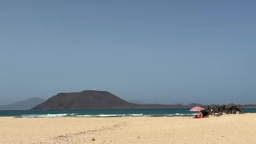 A wide shot of a sandy beach with a person sitting under a red and white striped umbrella. The person is partially visible, wearing a red garment. In the background, there is a body of water with waves and a dark, conical island or volcano in the distance. The sky is clear and blue. The sand is light brown and appears textured.