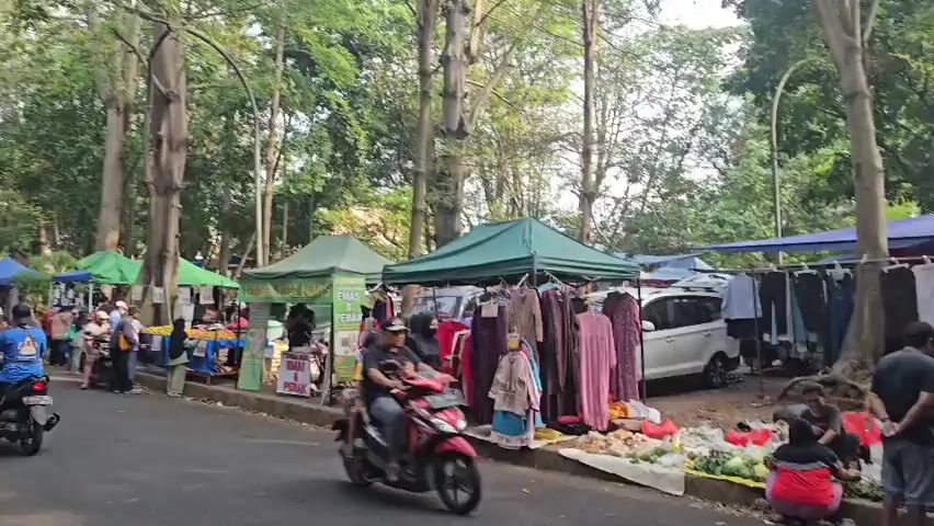 A street market is in full swing with numerous stalls set up along the side of a paved road. The market appears to be outdoors, under the shade of tall trees. Several vendors are displaying their goods, which include clothing, such as dresses and shirts, as well as produce like fruits and vegetables laid out on mats. People are seen browsing the stalls, walking along the road, and riding motorcycles. One stall has a sign that reads "PULSA & PAKET DATA" and another appears to advertise food. The atmosphere is lively and bustling, suggesting a typical day at a local market.