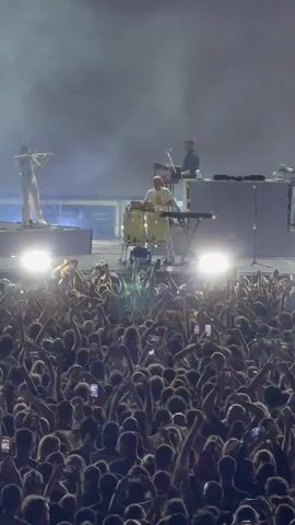 A concert performance unfolds with a drummer center stage, playing congas. To their left, a violinist stands, and to their right, another musician works at a console. Behind them, a large screen displays abstract blue and white graphics. Below the stage, a dense crowd of concertgoers raises their phones, their screens illuminated. Bright stage lights illuminate the performers and spill into the audience. The atmosphere is dark with streaks of smoke.
