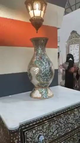 The video shows a piece of furniture, a chest of drawers, standing against a column decorated with horizontal stripes in orange, white, and gray.  The chest, which has a white marble top, is ornately decorated with inlaid mother-of-pearl. A vase, also decorated with mother-of-pearl, stands on top of the chest. An ornate lantern hangs above the vase. People walk in the background of what appears to be an indoor exhibition space, with national flags visible.