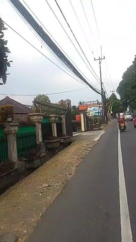 The video shows a street-level view of a road with overhead wires. On the left side of the road, there is a fence, a small drainage ditch, and some vegetation. In the background, there is a building with a sign that reads "HOTEL." Further down the road, there are more buildings and some flags. A motorcycle is visible on the right side of the road, moving away from the camera. The sky is overcast.