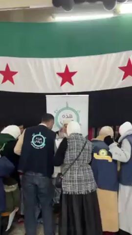 A group of people are gathered around a table, seemingly involved in some kind of registration or distribution process. Many are wearing blue jackets with a circular logo and Arabic writing on the back. Some individuals are wearing headscarves. A white banner with similar circular logo and Arabic script, reading "هذه حياتنا" (This is our life) in green, stands behind the table. The background features a flag with horizontal green, white, and black stripes and three red stars, resembling the Syrian flag. The scene appears to be taking place indoors.