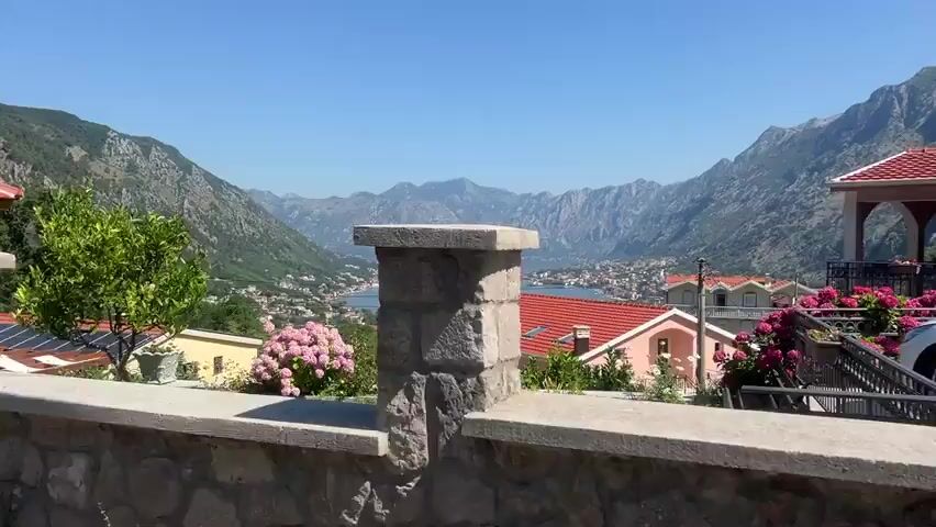 The video shows a scenic view of a bay with mountains in the background. There are several houses with red roofs visible, some with solar panels. In the foreground, there is a stone wall and a tree. The water in the bay is calm, with a few boats and a cruise ship. The sky is clear and blue. The video appears to be taken from an elevated position, looking down on the bay and the town. The overall impression is one of a peaceful and beautiful summer day in a coastal town.