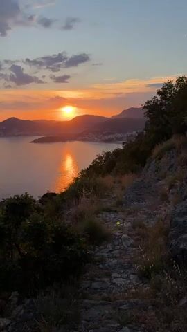 A vertical video captures a sunset over a body of water and distant hills. The sun, a bright orange orb, is low on the horizon, casting a warm glow and its reflection across the water. Wispy clouds are visible in the sky, painted with shades of orange and purple. The foreground features a rocky path winding along a hillside covered in vegetation, including trees and dry grass. The scene appears to be a scenic overlook, possibly a hiking trail, offering a panoramic view of the coastal landscape at dusk.