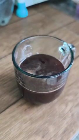 Hot water is being poured into a clear glass mug containing dark liquid, likely coffee grounds. The mug is sitting on a wooden surface. The water stream creates ripples and foam as it mixes with the coffee. The background is blurred but appears to be a kitchen counter with some objects.