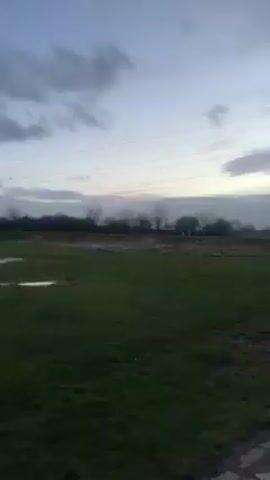 The video shows a grey, overcast day at a park. The camera starts focused on a patch of grass and a winding pathway before panning to the right, revealing two massive metal horse head sculptures, The Kelpies, in the distance. The sculptures appear silver in color, with a complex, geometric design. The sky is filled with dark clouds, and power lines are visible in the background. Someone is walking along the pathway towards the sculptures. The camera continues to pan across the sculptures, providing different angles of the horse heads against the backdrop of the cloudy sky and the surrounding park.