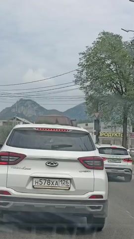 The video shows a white Chery Tiggo 4x4 SUV from behind, driving on a paved road. The license plate reads H578XA 126. Mountains are visible in the background. The sky is overcast. To the right of the road, there are trees, power lines, a traffic light, a street sign that says "ПРОДУКТЫ" (Products), and other vehicles. The SUV has its brake lights on. Smoke or dust is coming from under the rear bumper of the SUV.