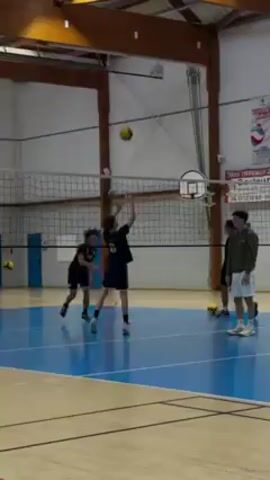 The indoor video shows a volleyball practice in a gymnasium. Six young men, dressed in athletic attire, are on the volleyball court. One player leaps into the air and spikes the volleyball over the net. The other players react to the spike, while some stand observing the game. A basketball hoop hangs from the ceiling above the net. Banners and posters adorn the wall. The floor is blue.