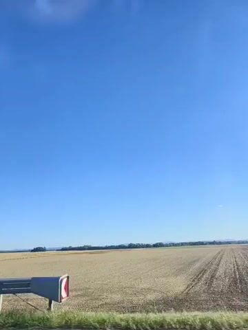 The video shows a view from a moving vehicle, likely a car or bus, traveling along a highway. The scene is dominated by a vast, clear blue sky and a golden field of harvested crops stretching to the horizon. A metal guardrail runs along the bottom of the frame, separating the road from the field. In the distance, a line of trees marks the edge of the field, and faint hills are visible beyond them. The sun appears to be shining brightly, casting a warm glow on the landscape. The overall impression is one of open space and rural scenery.