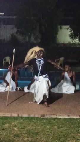 Here is a detailed description of the video:

The video captures an outdoor performance at night. The central figure is a man dressed in a black shirt, a white wrap-around garment, and a straw-like wig. He is holding a long, thin staff in his left hand, while his arms are outstretched to the sides. He appears to be actively performing or dancing, with an excited expression on his face.

Behind him, two individuals are seated on the edge of a swimming pool, each draped in white cloths. To the left of the main performer, a man sits cross-legged, holding a similar staff. To the right, a woman sits, and she looks directly at the main performer.

The performance area is a paved surface, possibly a patio or a stage, bordered by a grassy area. The background includes trees and a structure, potentially a building.

The lighting is artificial, creating visible shadows and indicating an evening setting. The overall atmosphere suggests a cultural performance or a staged event.