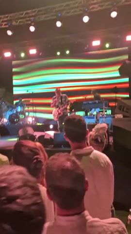 The video shows a concert with a band performing on a stage. A guitarist is in the center of the stage, wearing a colorful shirt. Behind him, a drummer and keyboardist can be seen. The stage is lit with colorful lights and a large screen displays a vibrant, abstract pattern. In front of the stage, a crowd of people is gathered, many holding up their phones to record the performance. The audience appears to be enjoying the show, with some people dancing and cheering. The lighting shifts between bright white, pink, red, and green hues, creating a dynamic atmosphere. The scene is dark except for the stage lights and the illuminated screens.
