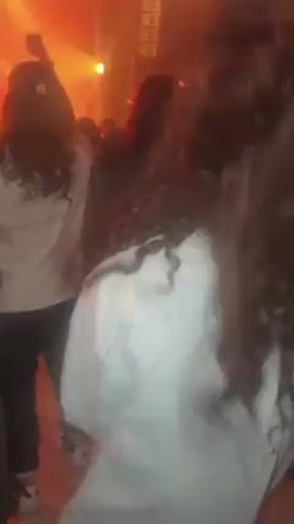 The video shows a crowd of people at an indoor event, possibly a concert or a rave. The lighting is primarily orange and red, and there is some smoke in the air, creating a hazy atmosphere. People are dressed in casual attire, with some wearing band shirts or jackets with logos on the back. Most of the crowd is facing away from the camera, making it difficult to discern individual faces. There are speakers on the wall. The camera moves slightly.