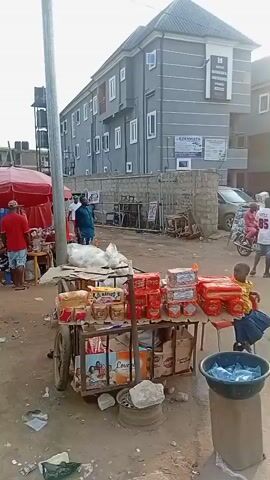The video shows a bustling street scene in what appears to be a market area. There are several people present, some standing and conversing, others sitting. A man leans against a silver van. There are numerous vehicles, including cars and a van. A cart filled with goods like packaged food items sits prominently in the foreground. A red umbrella covers a stall. A large advertisement billboard is visible with the words "DON'T LET FEAR BLOCK FORTUNE." Debris and litter are scattered on the ground. A portable toilet is visible in the background. Two men shake hands.