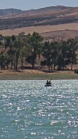 A wide shot shows a calm, glittering lake with a kayak in the middle distance. Two people are in the kayak, paddling. The lake is bordered by a dry, grassy shore with scattered trees. In the background, rolling hills are visible, with some agricultural fields and distant structures. The sky is clear and blue. The video shows the kayak moving gently across the water.