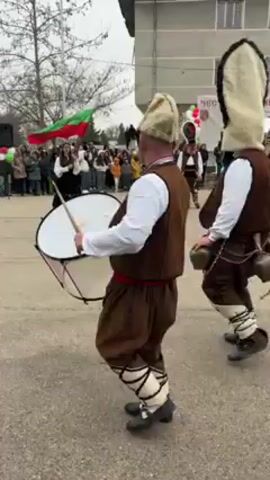 The video shows a group of people participating in a Bulgarian traditional festival, likely Kukeri. The focus is on several men wearing traditional costumes, including brown vests and trousers, white shirts, black shoes, and distinctive tall, white, conical hats. They also have belts with bells attached, which jingle as they walk. One man beats a drum, adding to the festive atmosphere. The group parades down a street, passing a crowd of onlookers and a building with the text "ЧЕСТИТ ПРАЗНИК АБЛАНИЦА" visible on it. A Bulgarian flag is also visible in the background. The environment appears to be a small town or village, and the weather seems overcast.
