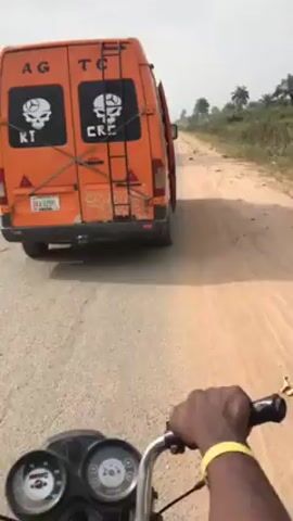 A first-person perspective video shot from a motorcycle shows the driver following an orange van on a road. The van has two skull logos on the back, each with text below. The road the vehicles are on is paved, but an adjacent dirt road can be seen alongside it. The environment appears rural, with trees and vegetation visible in the background. The sky is overcast. The motorcycle driver's hand can be seen on the handlebar.