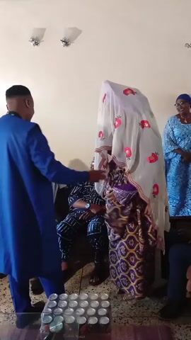 A man in a blue outfit stands to the left, facing a woman covered in a sheer white veil with red floral patterns. He is pulling the veil away from her face. Two men are seated on a couch behind the woman. One man is wearing a grey outfit and a red hat, and the other is wearing a blue and white patterned outfit. Another person is visible on the far right, and a woman is partially visible on the far left. The room appears to be a living room with light-colored walls and two wall-mounted lights. In the foreground, there is a glass table with bottles and a tray of small metallic cups. The floor is tiled.