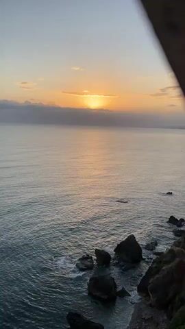 The video shows a sunset over the ocean, with a rocky coastline in the foreground. The sun is low on the horizon, partially obscured by clouds, and its light reflects on the water. Waves crash against the rocks, and a bird flies across the scene. The overall impression is one of natural beauty and tranquility.