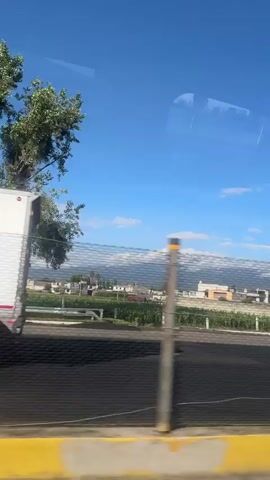 This is a vertical, first-person perspective video taken from a moving vehicle, likely a car or bus, looking out a window. The view is of a highway with other vehicles and a landscape in the background. The sky is clear blue with a few scattered white clouds. On the right side of the road, there is a large white truck with a cargo box filled with yellow crates. To its left, there is a large red shipping container with the "MSC" logo on it, being transported on a trailer. Further to the left, there is a silver car. The road has a yellow barrier on the immediate right. In the distance, there are buildings, fields, and hills. There are also some power lines and poles visible. The video appears to be taken during the daytime.