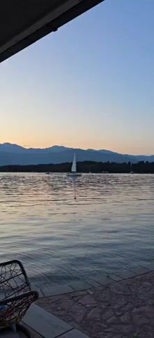 The video shows a sailboat on a body of water. The sun is setting, and the sky is a mix of orange and blue. In the background, there are mountains. The water is calm with small ripples. There are other boats in the distance.