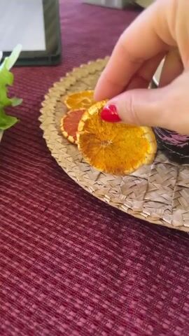 The video shows a close-up view of a person's hand with red nail polish picking up a dried orange slice from a woven mat. The orange slice is golden brown and appears to be a garnish. The hand then dips the orange slice into a white teacup filled with a light brown liquid. Another dried orange slice and a dried pink grapefruit slice are visible on the woven mat next to the teacup. The background is blurred, but a pink tablecloth and a green leafy object are partially visible. The lighting suggests an indoor setting.
