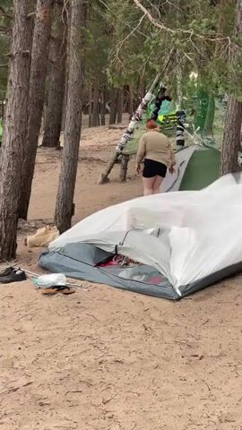 A sandy beach is seen with several tents set up among trees. A person in a white t-shirt and dark shorts walks across the sand, passing by a table and a grill. In the foreground, a partially collapsed tent is visible, along with some shoes and a mask on the sand. In the background, another person wearing a light-colored jacket and dark shorts is seen walking away from the camera. Several other tents and camping gear are scattered around the area. The trees are dense, and the ground is covered in sand and pine needles. The overall scene depicts a campsite on a beach.