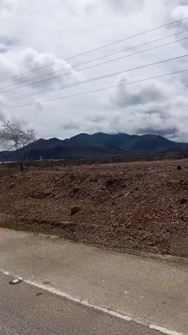 The video shows a roadside view of a dry, arid landscape. In the foreground, a paved road with a white line is visible. Beyond the road, the ground is covered in dry soil, small rocks, and sparse, dry vegetation. A solitary, bare tree stands in the middle ground. In the background, a mountain range is visible under a cloudy sky. Power lines and a utility pole are also present in the scene. The camera appears to be in motion, suggesting a drive-by perspective. The overall impression is of a remote, undeveloped, and dry environment.