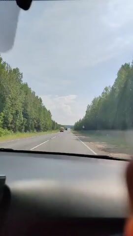 The video shows a POV of someone driving on a highway. The road is asphalt and has two lanes in each direction, separated by a white line. The road is surrounded by dense green trees on both sides. The sky is blue with scattered white clouds. Several vehicles are visible on the road, including cars and trucks. One truck is passing the vehicle the video is being filmed from. The lighting indicates it is daytime, likely sunny. The interior of a car, specifically the dashboard, is visible in the foreground.