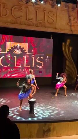 In an indoor venue, likely a hotel ballroom or theater, several female dancers perform on a stage. The stage is lit with spotlights creating circular patterns on the floor. A large LED screen behind the performers displays a logo that reads "BELLIS DELUXE HOTEL". To the right of the stage, a large golden, feather-like sculpture is visible. The dancers are dressed in colorful, sparkly costumes. One dancer in the foreground, wearing a blue and silver outfit, stands on top of a djembe drum. She is performing a dynamic dance routine, at times balancing on one leg, at other times bending over. Two other dancers in similar attire are also on stage, performing synchronized moves. Another dancer, in a pink and silver outfit, is further back on the right side of the stage, also executing dance steps. The overall atmosphere appears to be a lively performance.