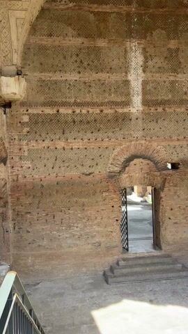The video shows the interior of an ancient structure. The walls are constructed from brick and a rough, textured material, with visible patterns and layers. There are arched doorways, one of which has a metal gate. The ceiling is vaulted and features decorative patterns, though it appears aged and deteriorated. The light indicates it is daytime. There is a glimpse of a staircase and a railing in the foreground.