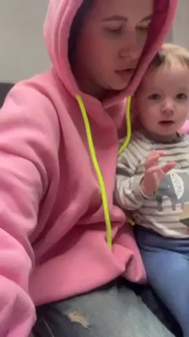 A person wearing a pink hoodie with yellow drawstrings is holding a young child wearing a long-sleeved striped shirt with an elephant graphic. The person in the hoodie is looking down at something in their hands and appears to be reading or talking. The child is looking down at their hands and touching something in front of them. The background is a blurry, undecorated wall.
