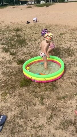 This video captures a summer day at a sandy beach in Vangaži, Latvia. The primary subjects are two young children playing near a small, inflatable swimming pool. One child, wearing a diaper and a hat with a tiger print, is standing in the shallow water of the pool. Another child, also wearing a diaper and a patterned headscarf, is perched on the edge of the pool behind the first child, reaching down towards the water.

The scene is set on a sandy area dotted with dry grass and patches of green vegetation. In the background, more sand can be seen, leading up to a faint outline of a volleyball net and some buildings, suggesting a recreational area. A few items of clothing are visible on the sand in the distance. The lighting suggests it is daytime, likely sunny and warm, suitable for outdoor water play. The children appear to be engrossed in their play, with no strong emotional cues visible. The overall atmosphere is one of casual summer fun.
