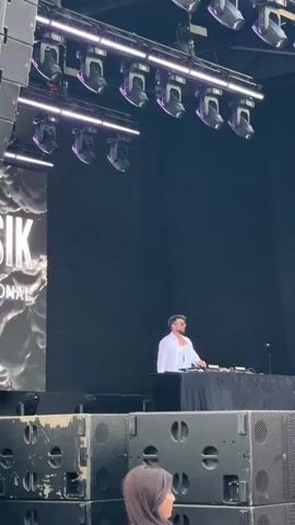 A male DJ in a white shirt and sunglasses stands behind a DJ booth on a stage. He is mixing music, gesturing with his hands as he works. The stage is equipped with professional sound and lighting equipment, with large speakers visible in the foreground. A large screen displays abstract, colorful imagery. The overall setting suggests an outdoor music event or festival.