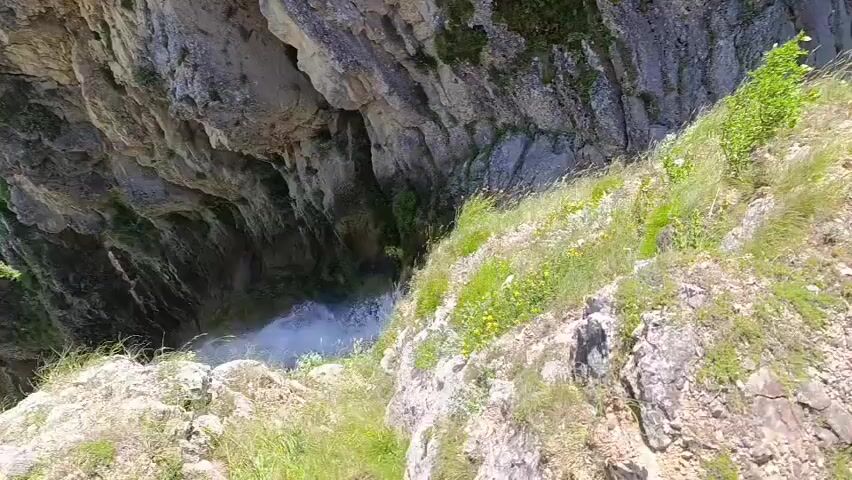 The video pans over a lush green gorge with a waterfall cascading down a cliff face. The camera starts from the edge of the cliff, looking down at the waterfall and the gorge below. As the camera moves, it reveals the steep, rocky walls of the gorge covered in dense vegetation. The waterfall is visible in the center of the frame, with its spray creating a misty effect. The scene is bathed in bright sunlight, indicating it is daytime. The overall impression is one of a remote and natural landscape.