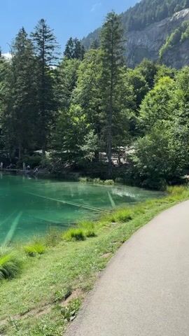 The video shows a scenic view of a clear, turquoise lake surrounded by lush green trees and a majestic mountain range in the background. The sun is shining brightly, casting dappled light on the water and the surrounding vegetation. Several people can be seen enjoying the scenery, some walking along a paved path, while others are seated on benches or standing near the water's edge. There are submerged logs visible in the clear water, adding to the natural beauty of the scene. A young woman wearing a hat and a scout-themed scarf stands on a wooden bench, looking towards the lake. The overall atmosphere is peaceful and serene, with a focus on the natural beauty of the location and the leisurely activities of the visitors.