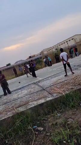 A group of about fifteen people, mostly young adults, are standing and walking on what appears to be a cracked cement sports court. They are observed in a casual outdoor setting, surrounded by grassy areas and buildings in the background. The weather looks overcast or it is late afternoon. Some individuals are wearing athletic apparel or casual wear, while others appear to be more formally dressed, possibly as part of a performance or event. There does not appear to be any specific activity taking place apart from the group's presence and movement within the court area. The camera is stationary and remains at a distance, capturing the general scene without focusing on any particular individual or action.
