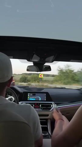 The video is filmed from the backseat of a car, looking forward through the windshield. In the foreground, two people are visible. The person in the driver's seat is wearing a white baseball cap and a white t-shirt. Their hands are on the steering wheel. To their right, a person is sitting in the passenger seat. Their legs are crossed, and they are wearing a dark top. Their right arm is extended, and their hand is resting on the arm of the driver. The driver's left hand reaches across and their fingers are intertwined with the passenger's hand. Both people appear to be looking forward, towards the road. The car's dashboard is visible, with a navigation screen displaying a map. The car's interior lighting is illuminated with purple accents. Outside the car, a road is visible, with trees and fields on either side. The sky is overcast. A yellow directional sign is visible on the right side of the road. Another car is visible in the distance, ahead of them.