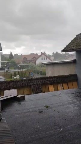 The video shows a view from a rooftop during a rainstorm. The sky is overcast with gray clouds. In the background, there are houses with red tile roofs and some trees. A fence separates a yard with children's playground equipment from the houses. In the foreground, the rooftop is wet with puddles of water. Some building materials, possibly tiles and wooden planks, are stacked on the rooftop. The rain is visible as streaks falling from the sky.